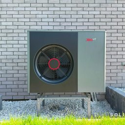 Solitar Fabryka Domów - Pompa ciepła JBG HT zamontowana na zewnątrz budynku z szarej cegły, otoczona białym żwirem i zieloną trawą. Widoczny fragment fundamentu i instalacja wodna.