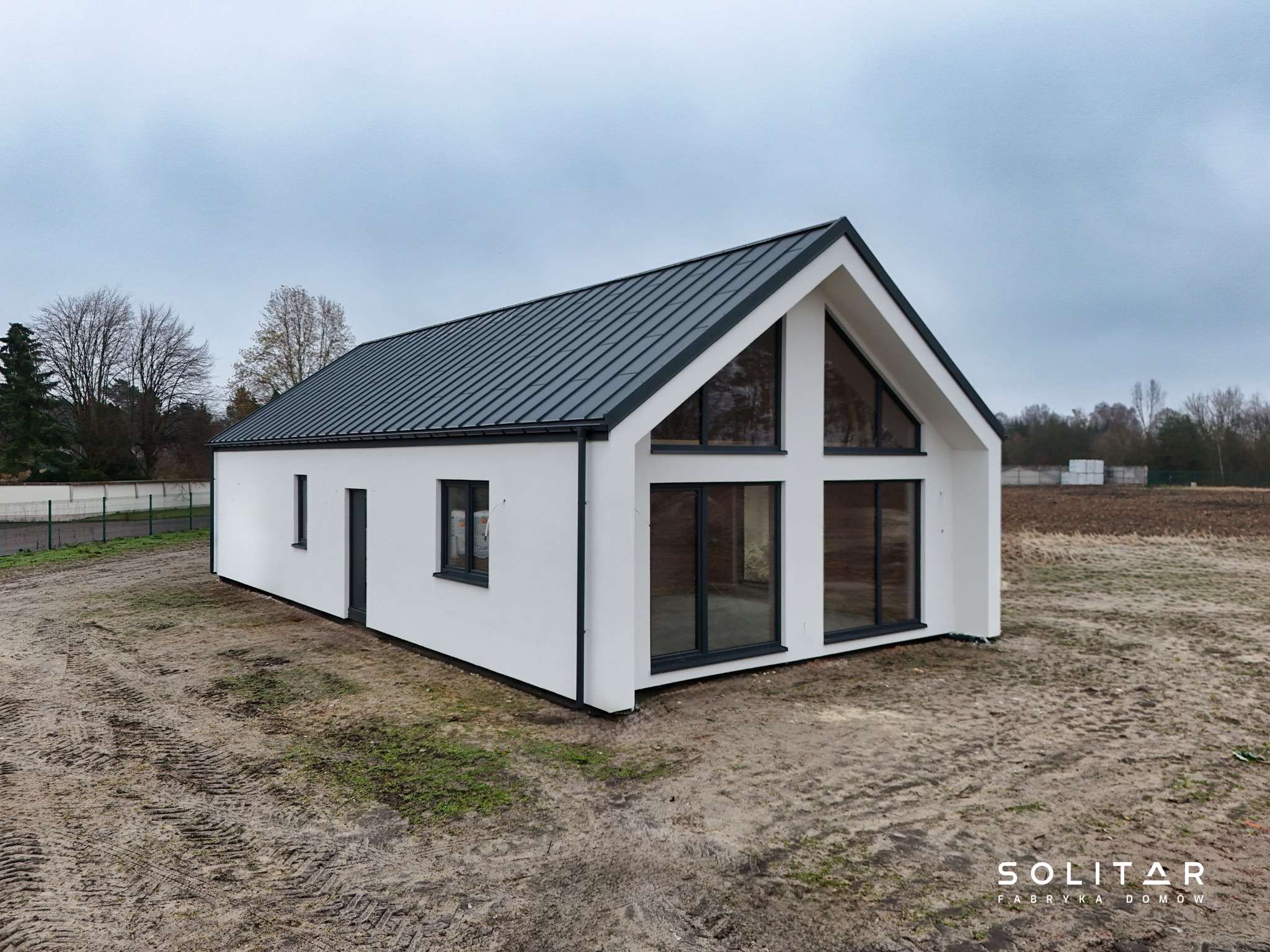 Nowoczesny, parterowy dom z białymi ścianami i grafitowym dachem na działce z widocznymi śladami opon. Duże okna z czarnymi ramami.