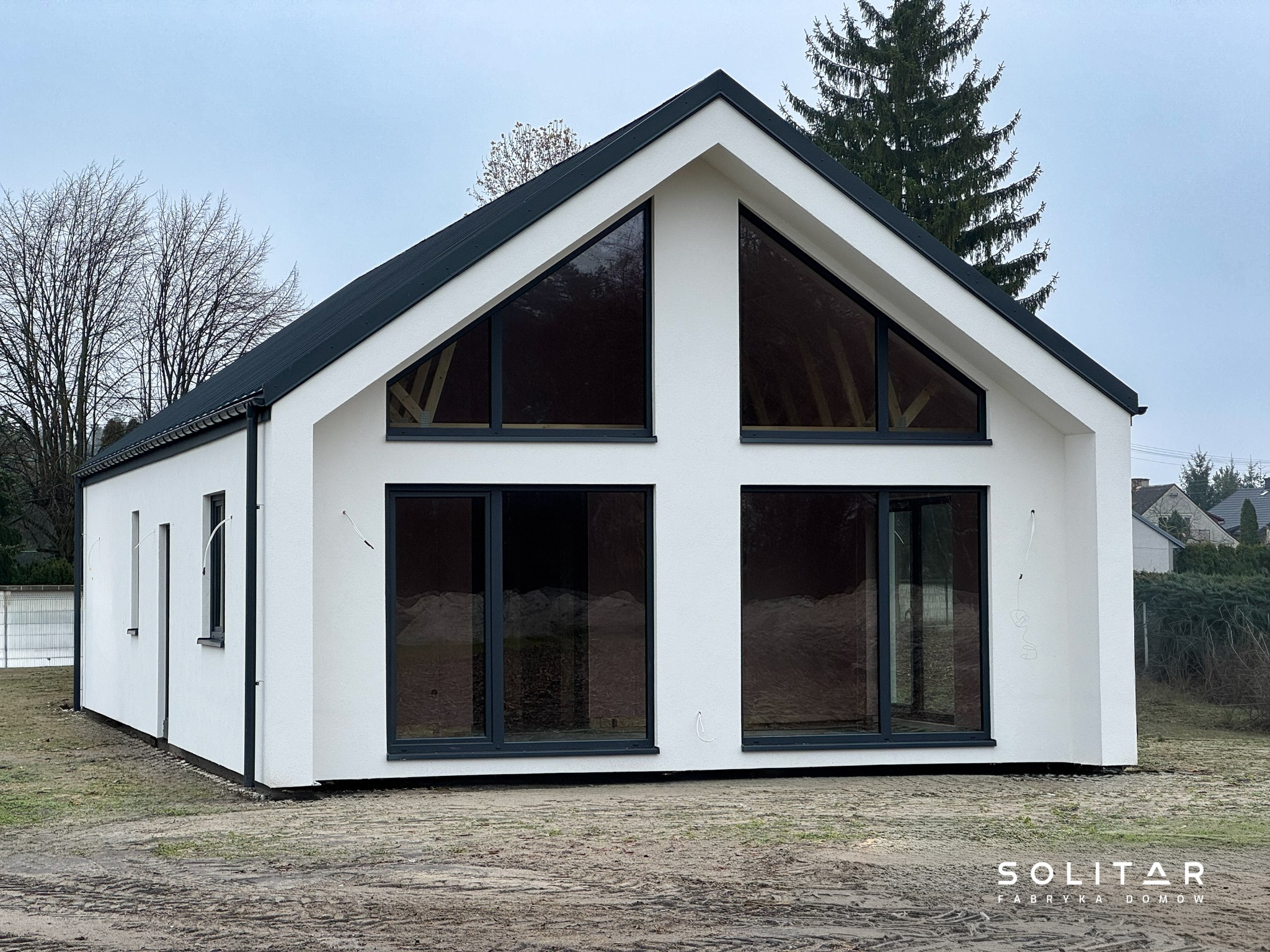 Biały, parterowy dom z czarnym dachem i dużymi oknami, widoczny z zewnątrz na tle drzew i pochmurnego nieba. Nowoczesna architektura z minimalistycznym designem.