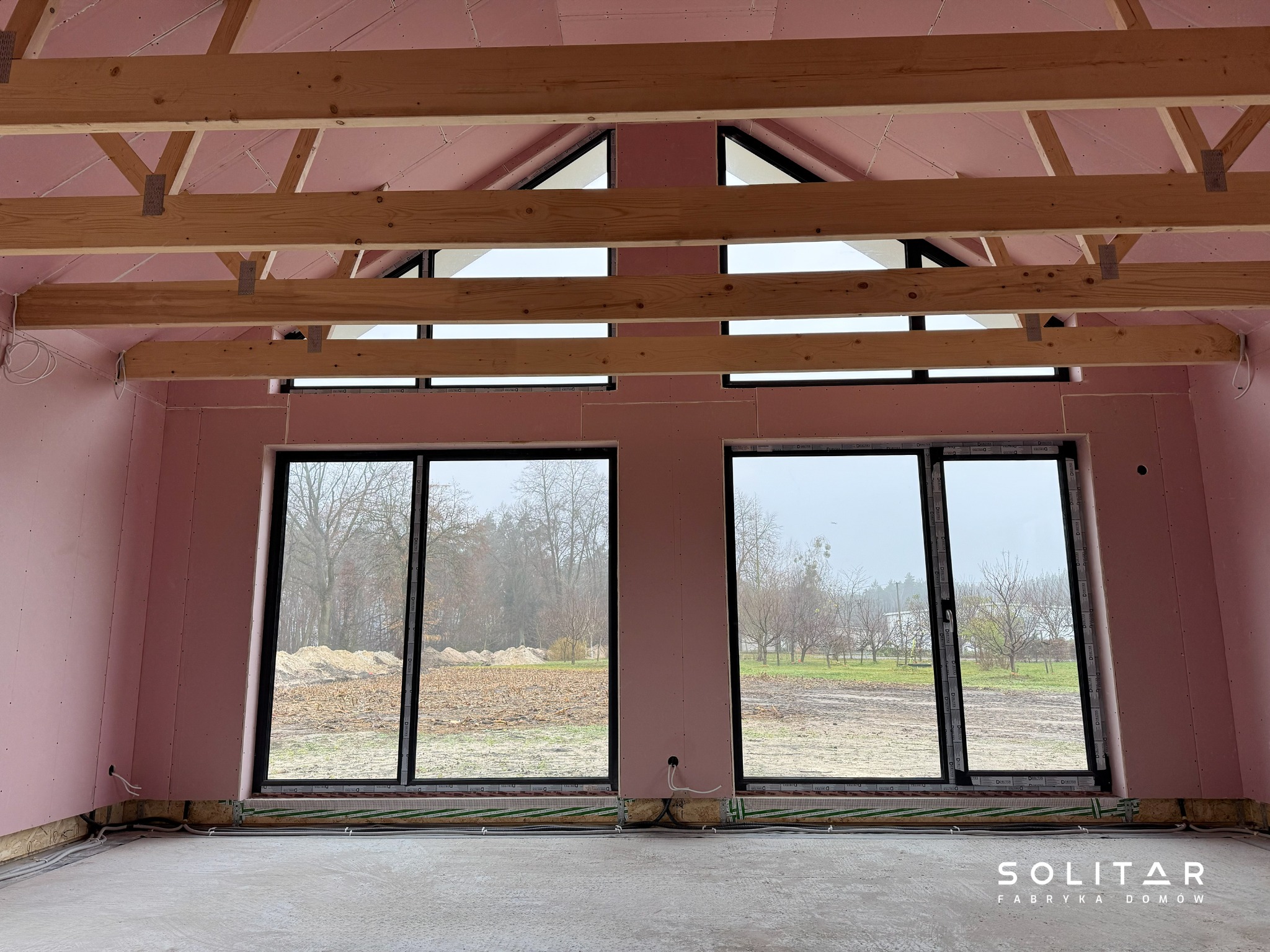 Wnętrze domu w trakcie budowy: różowe ściany, drewniana konstrukcja dachu, duże okna z widokiem na zewnątrz. Widoczne elementy instalacji podłogowej.