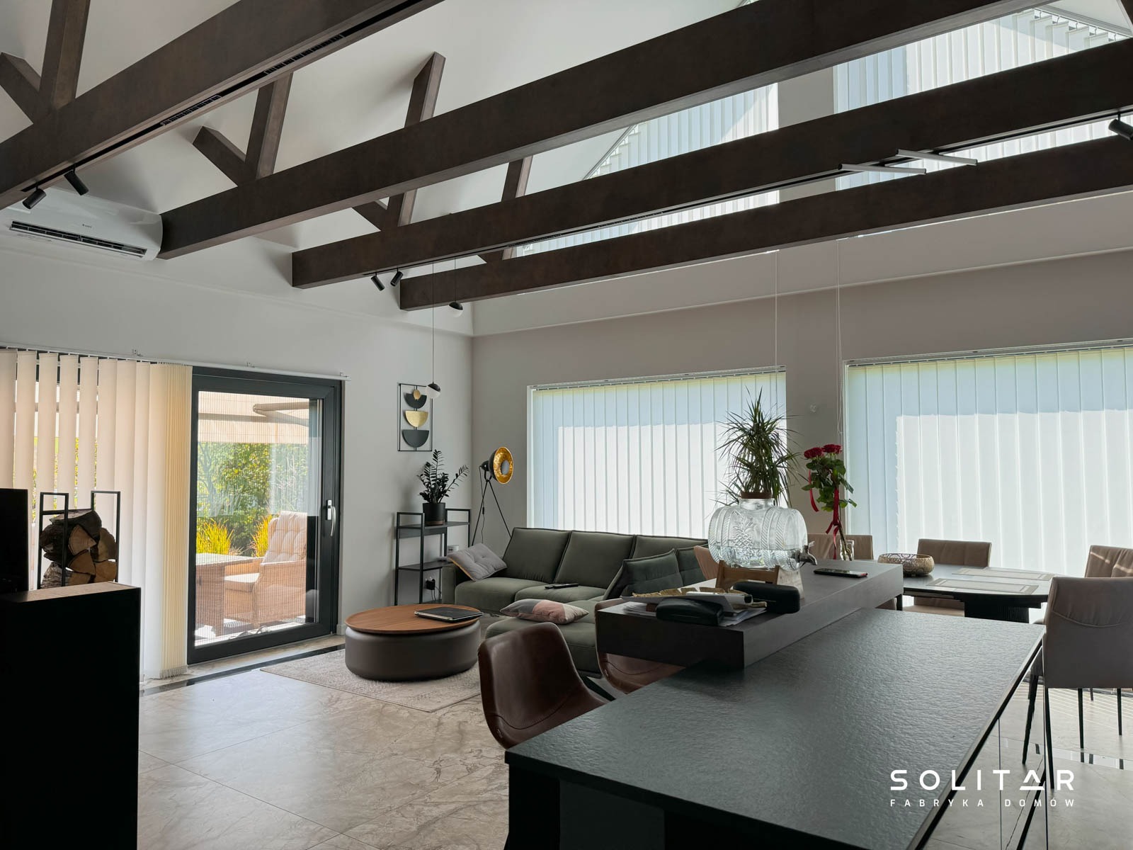 Jasne wnętrze domu z drewnianą konstrukcją stropu, dużymi oknami z żaluzjami pionowymi i nowoczesnym wyposażeniem. Widoczny salon z sofą i wyjściem na taras.