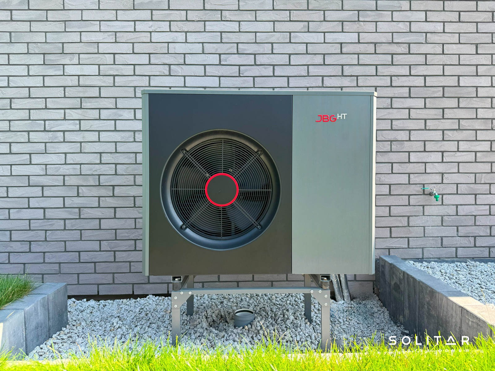 Pompa ciepła JBG HT zamontowana na zewnątrz budynku z szarej cegły, otoczona białym żwirem i zieloną trawą. Widoczny fragment fundamentu i instalacja wodna.