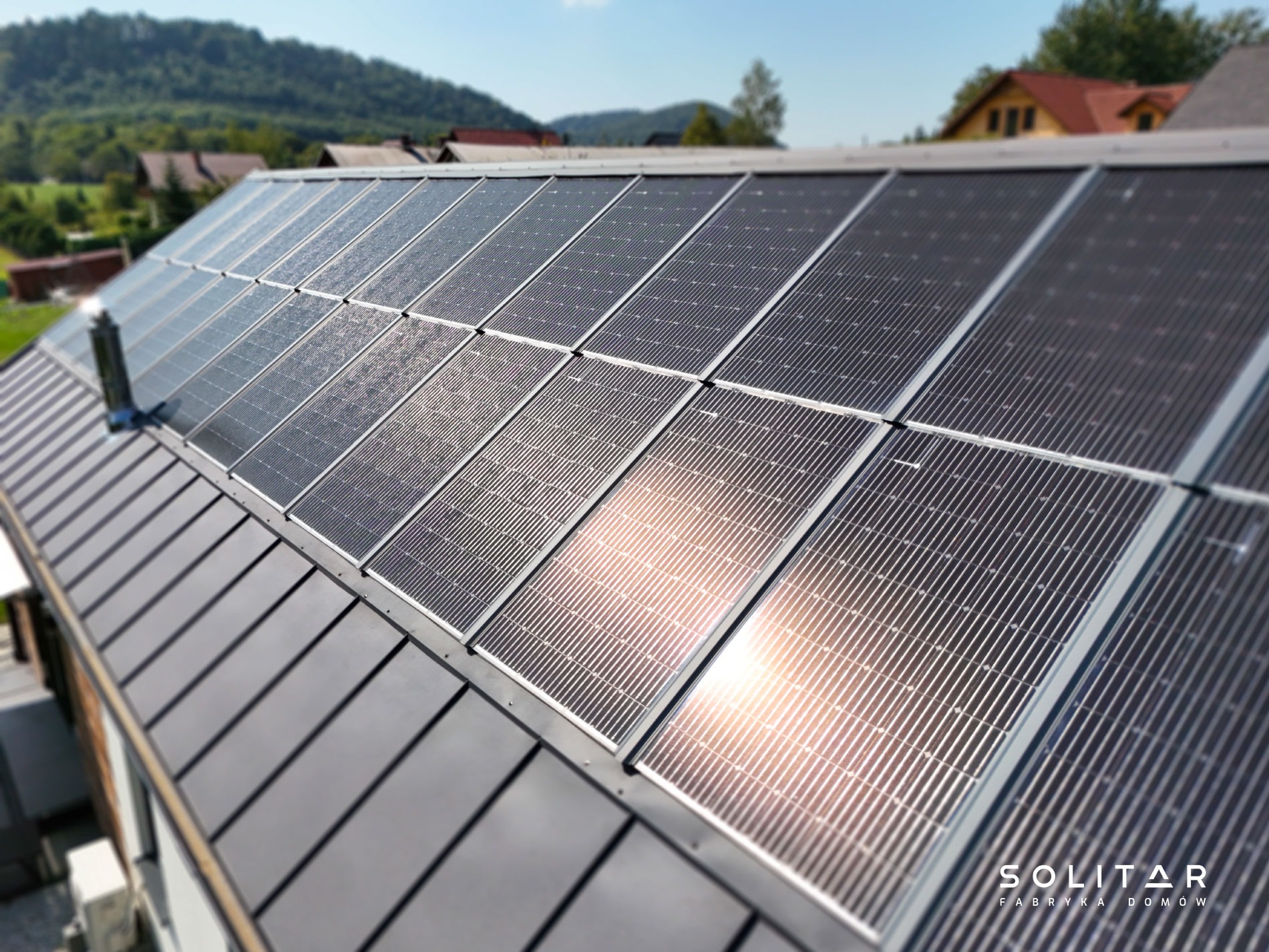 Panele słoneczne na dachu domu z szarej blachy, w tle zielone wzgórza i błękitne niebo. Widoczny komin i logo firmy Solitar Fabryka Domów.