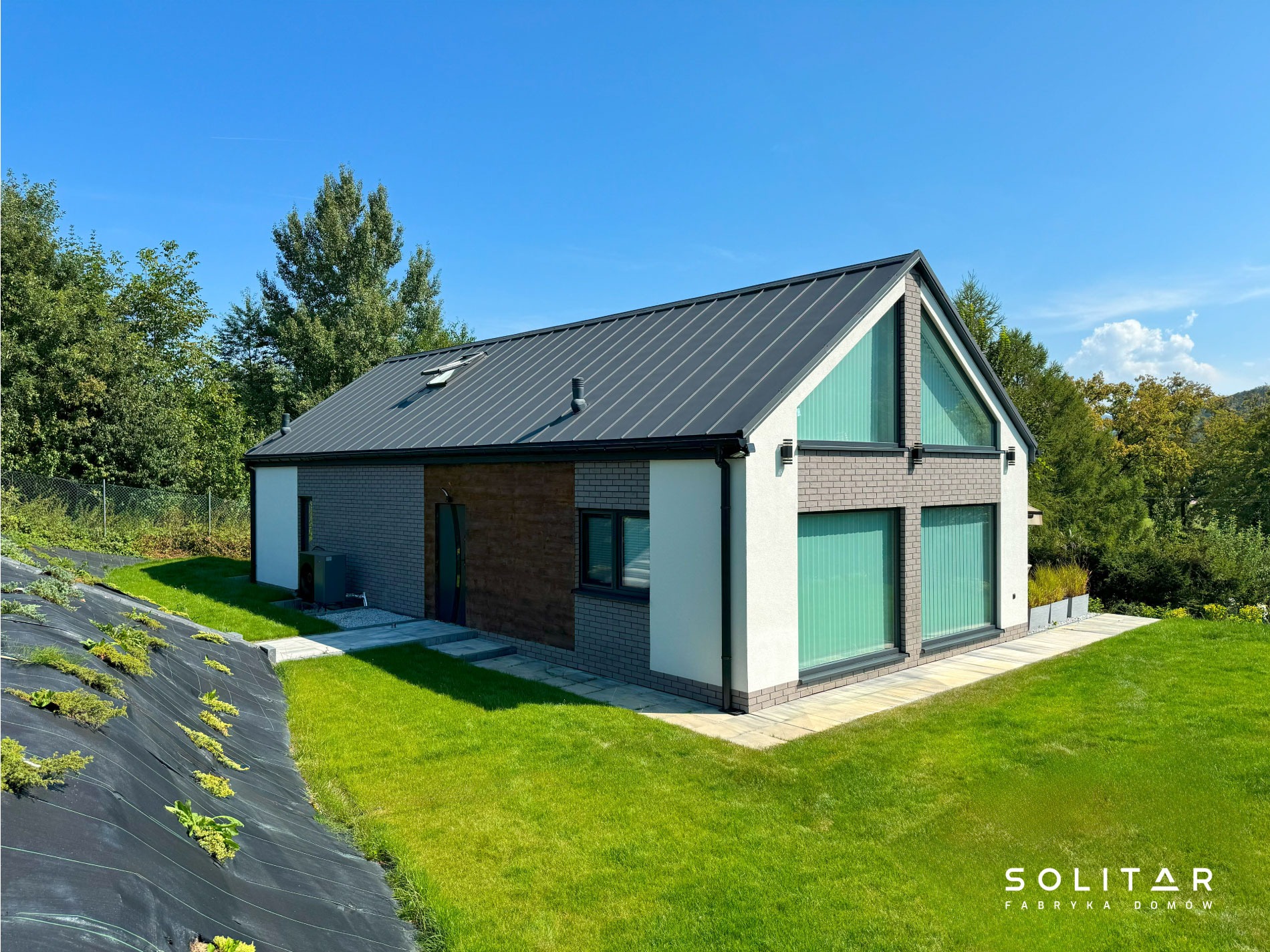 Nowoczesny, parterowy dom z szarą, metalową dachówką i dużymi oknami, otoczony zielonym trawnikiem i drzewami. Widoczny fragment ogrodu z roślinami okrywowymi.