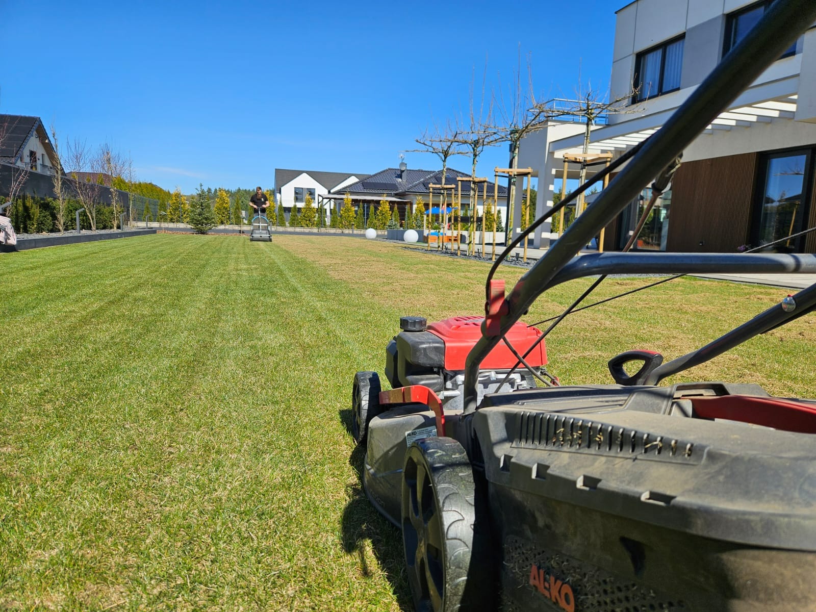 Częściowo skoszone trawnik przed nowoczesnym domem, widoczna kosiarka AL-KO na pierwszym planie i pracownik z kosiarką samojezdną w tle.