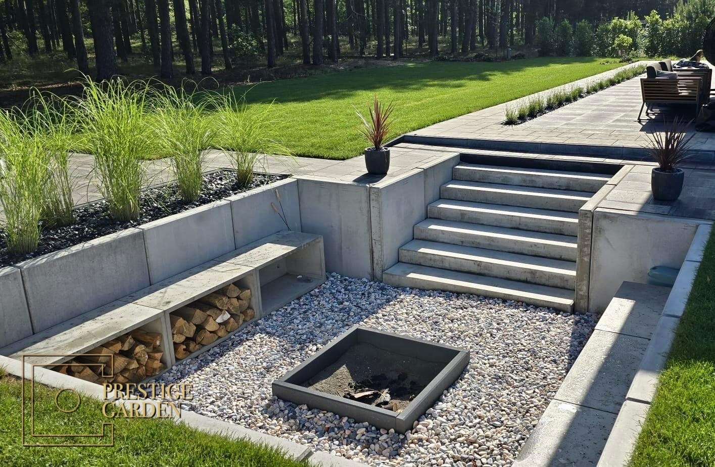 Nowoczesny ogród z paleniskiem otoczonym białymi kamykami, betonowymi schodami prowadzącymi na wyższy poziom z trawnikiem i drzewami w tle, oraz wnęką na drewno pod betonową ławką.