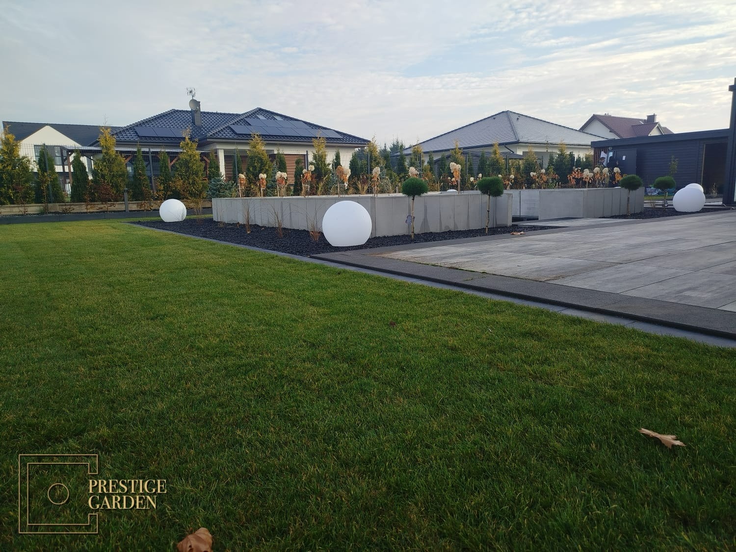 Nowoczesny ogród w Bydgoszczy z równo przystrzyżonym trawnikiem, podwyższonymi rabatami wykończonymi czarnym grysem, kulistymi lampami ogrodowymi i zadbanymi drzewami iglastymi.