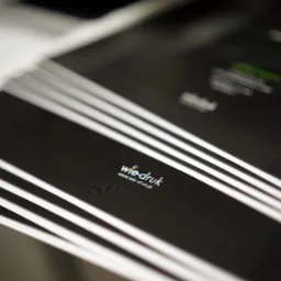 Świeżo wydrukowane, czarne arkusze z logo firmy drukarskiej 'wie-druk' i adresem strony internetowej, ułożone jeden na drugim w stosie.