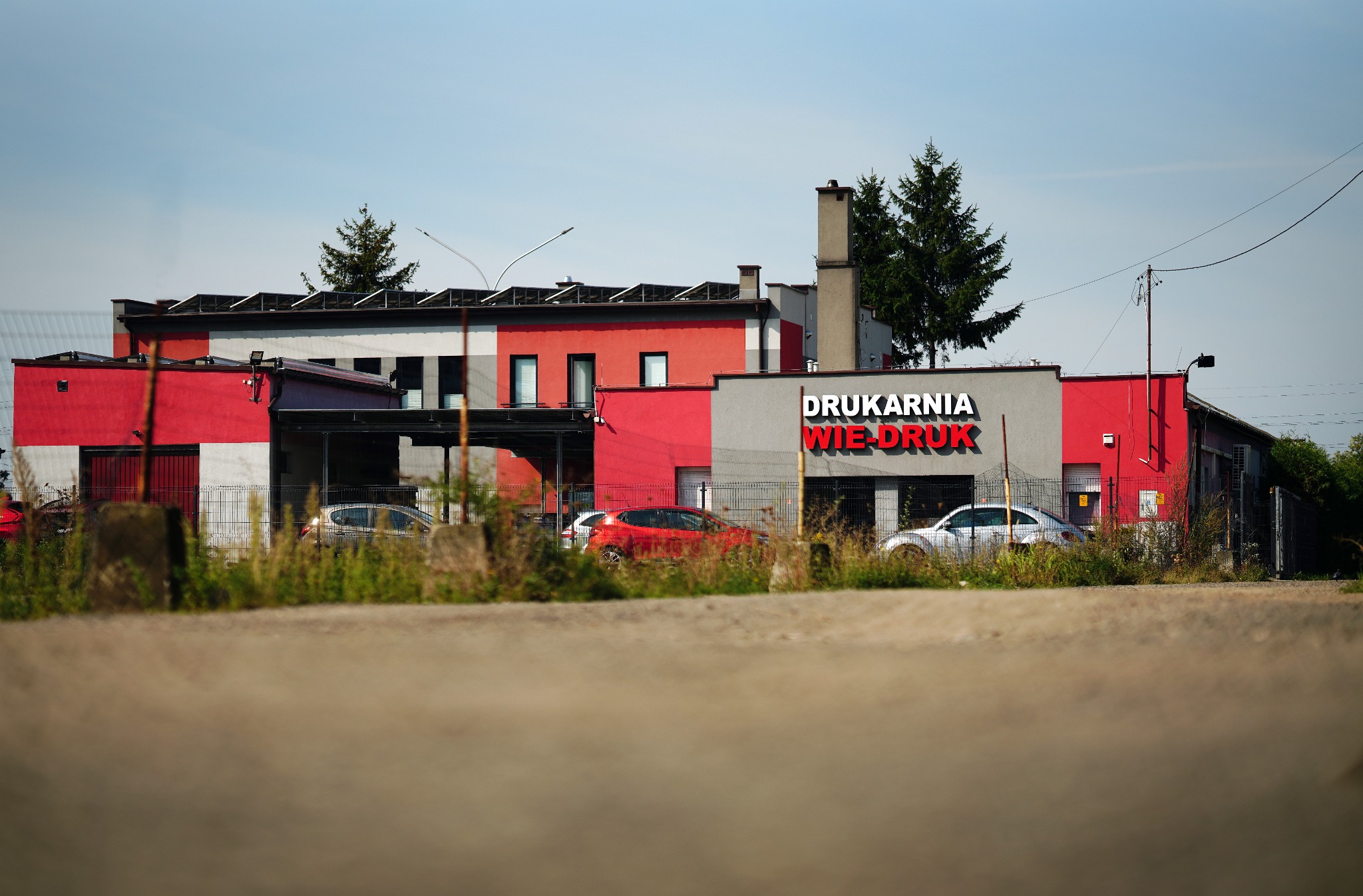 Budynek drukarni z napisem 'DRUKARNIA WIE-DRUK' na szarej fasadzie, czerwone i szare ściany, widoczne samochody i zieleń, ujęcie z niskiej perspektywy.