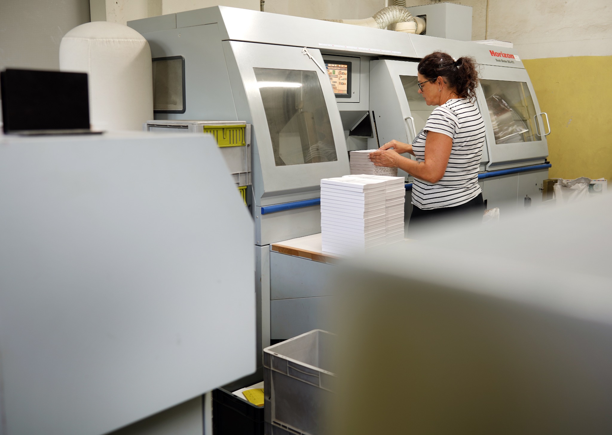 Pracownica w okularach obsługuje maszynę drukarską Horizon, układając stos białych arkuszy papieru; widoczny ekran sterowania maszyny i pojemnik na odpady.