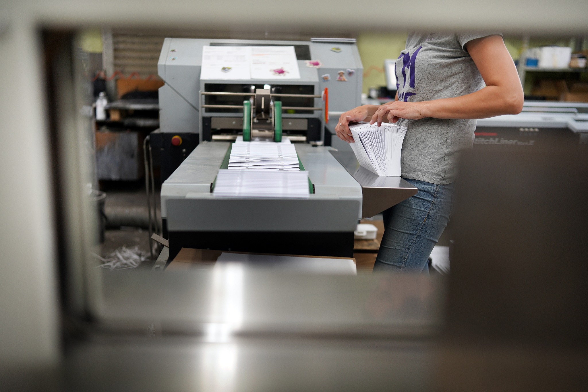 Kobieta w szarej koszulce i jeansach obsługuje maszynę do obróbki papieru, układając stos białych arkuszy na podajniku.