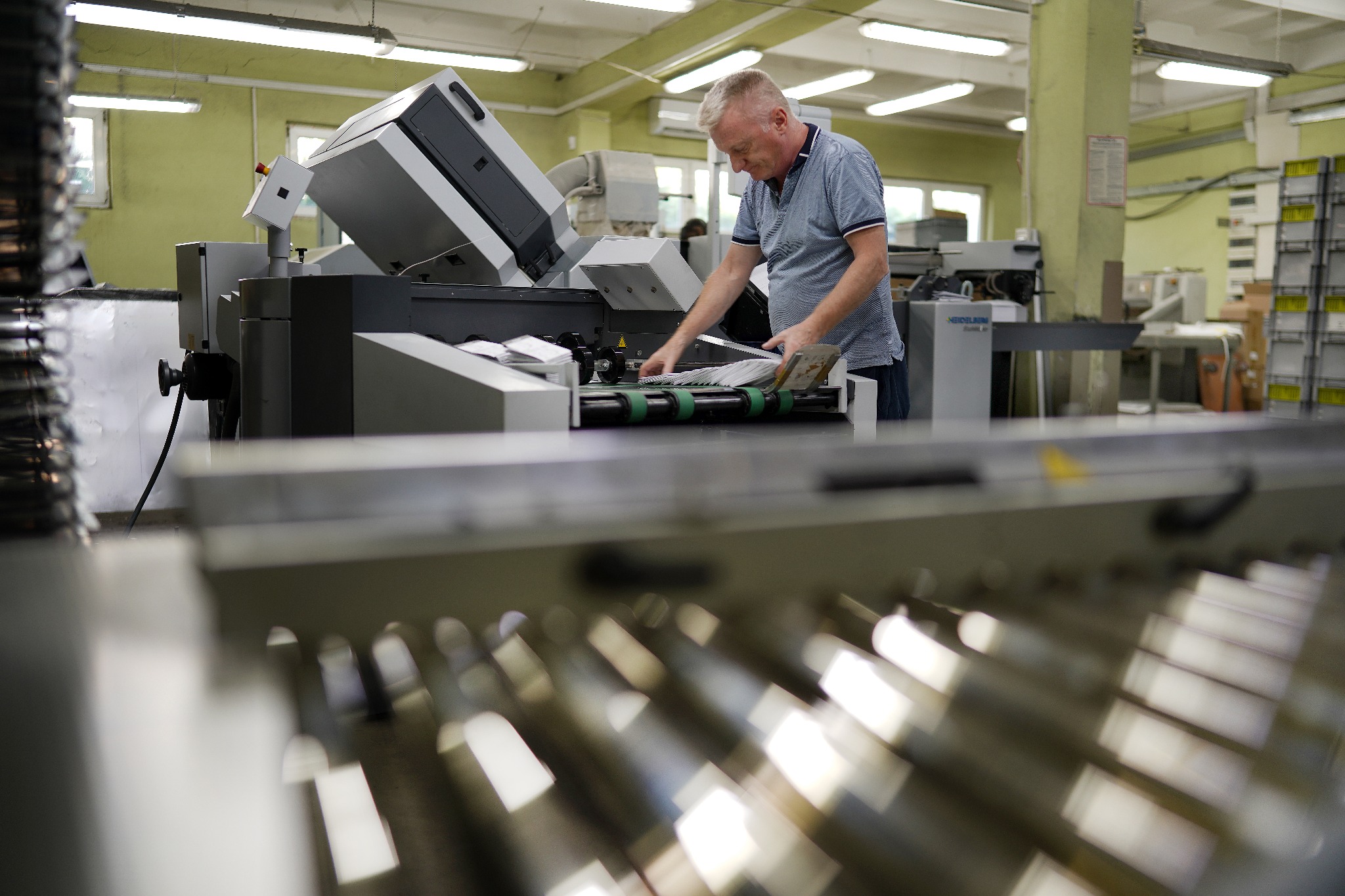 Pracownik drukarni obsługuje maszynę drukarską, układając arkusze papieru na podajniku. Widoczne elementy maszyny, oświetlenie przemysłowe i fragment hali produkcyjnej.