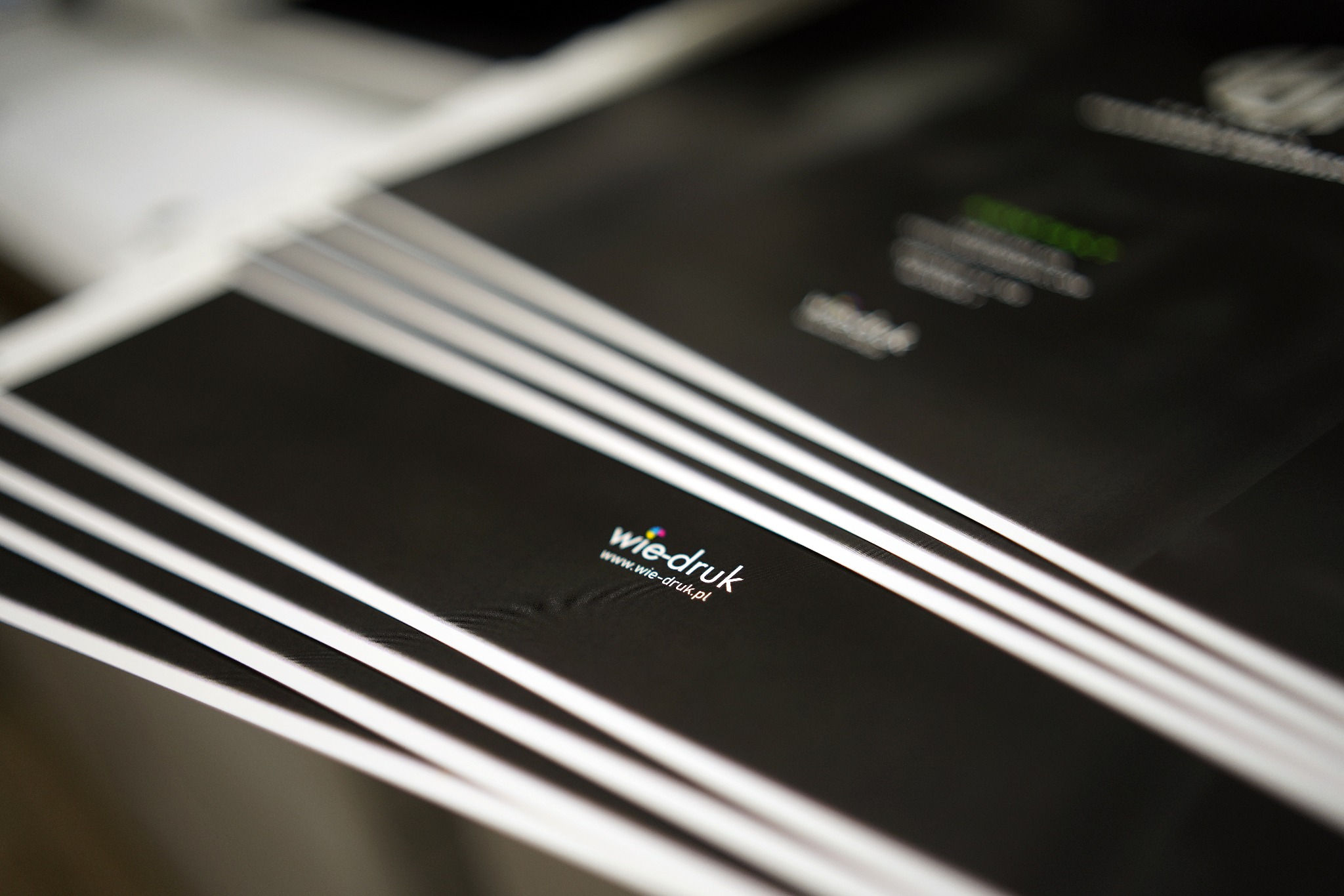 Świeżo wydrukowane, czarne arkusze z logo firmy drukarskiej 'wie-druk' i adresem strony internetowej, ułożone jeden na drugim w stosie.