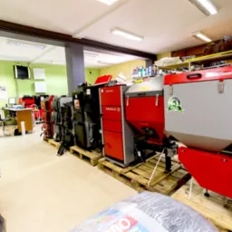 Wnętrze sklepu lub magazynu z kotłami grzewczymi różnych marek, w tym Defro Sigma 5 i Taurus, ustawionymi na paletach, widoczne również opakowania akcesoriów grzewczych i biurko w tle.