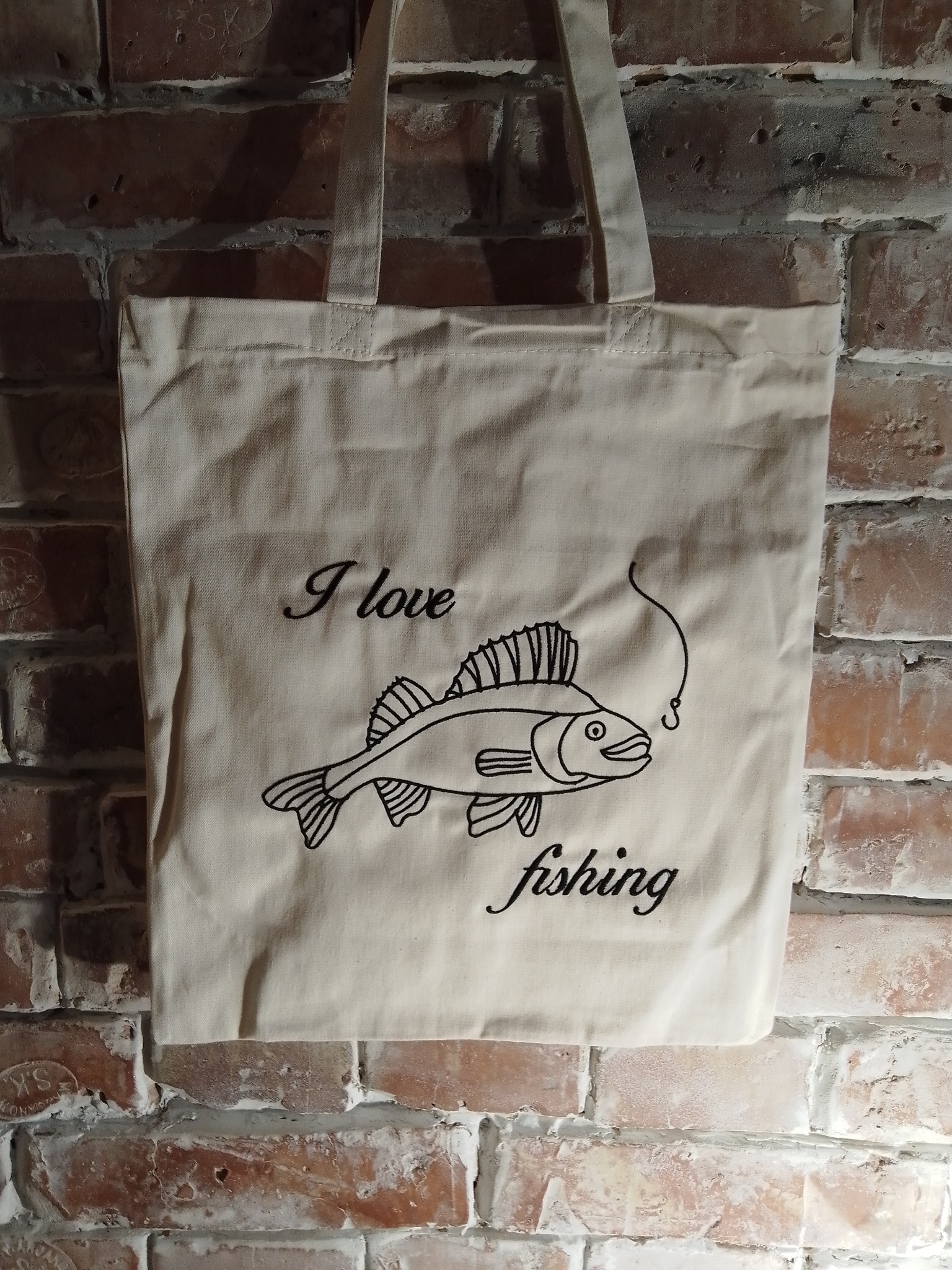 Beżowa torba bawełniana z czarnym haftem przedstawiającym rybę na haczyku i napisem 'I love fishing', wisząca na ścianie z czerwonej cegły.