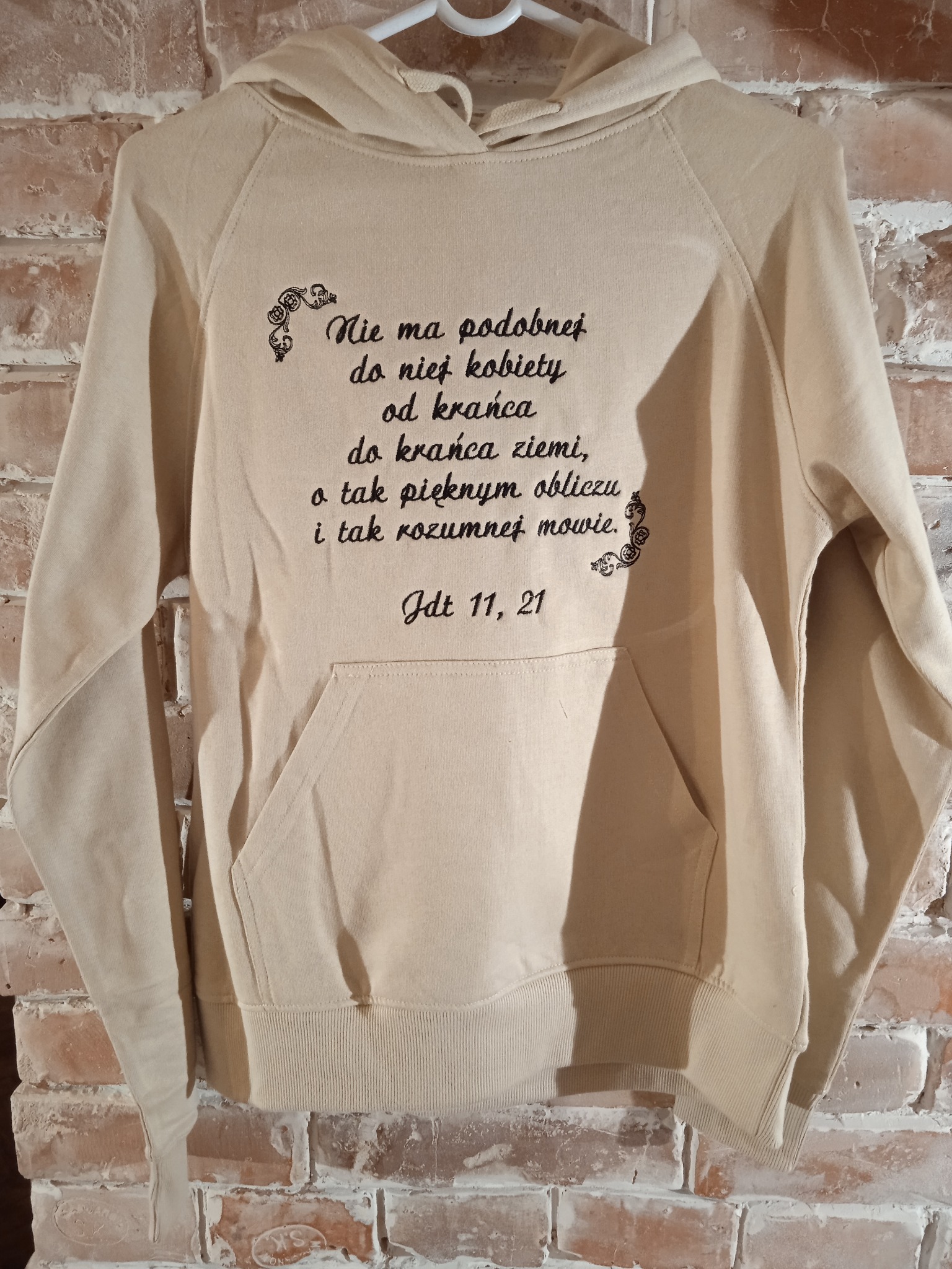 Beżowa bluza z kapturem, wisząca na wieszaku na tle ceglanej ściany, z wyhaftowanym czarnym tekstem 'Nie ma podobnej do niej kobiety od krańca do krańca ziemi, o tak pięknym obliczu i tak rozumnej...