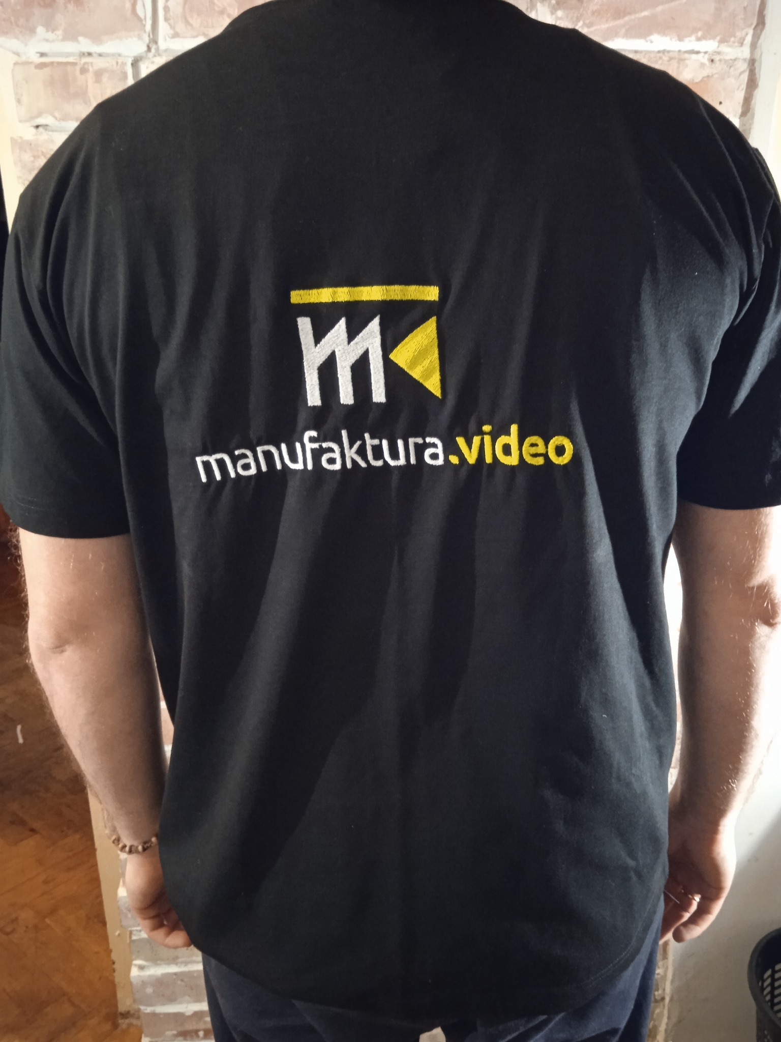 Czarna koszulka z haftowanym logo 'manufaktura.video' z tyłu, widok zza pleców mężczyzny.