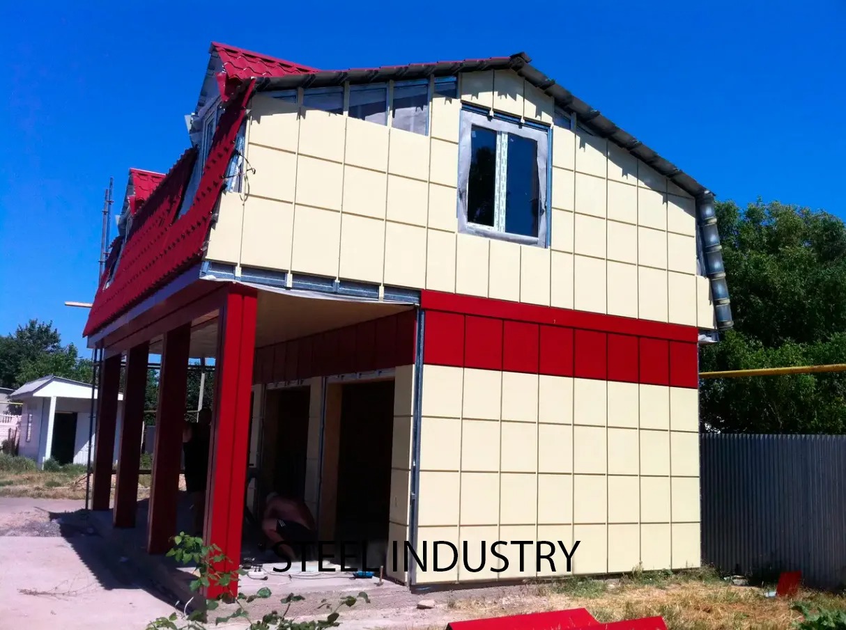 Dwukondygnacyjny budynek z czerwonym dachem i beżowo-czerwoną elewacją w trakcie montażu, widoczne metalowe profile i pracownik przy wejściu.