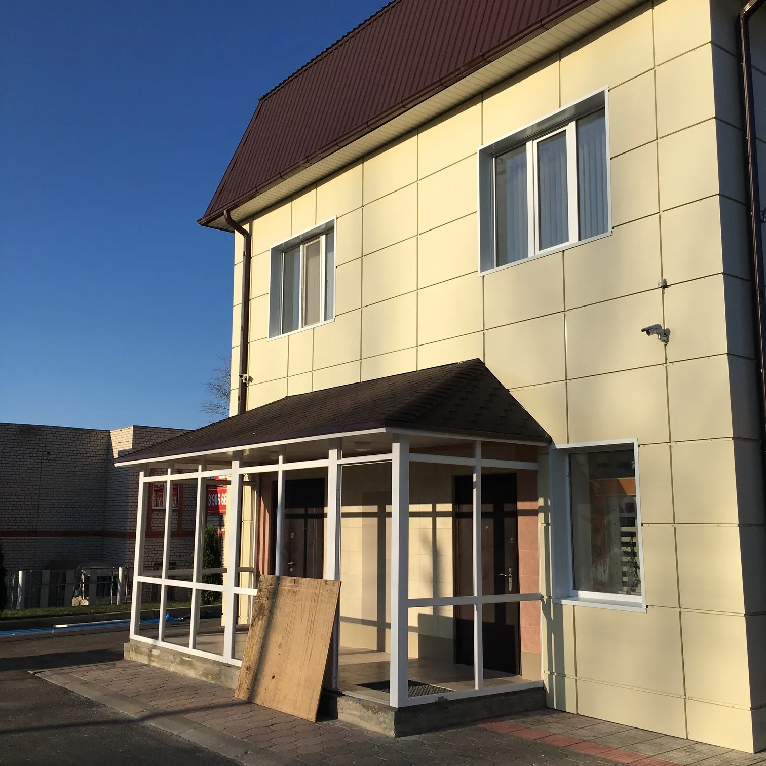 Budynek z elewacją z paneli w odcieniach beżu, z brązowym dachem i dobudowanym zadaszeniem nad wejściem. Widoczna kamera monitoringu na elewacji.