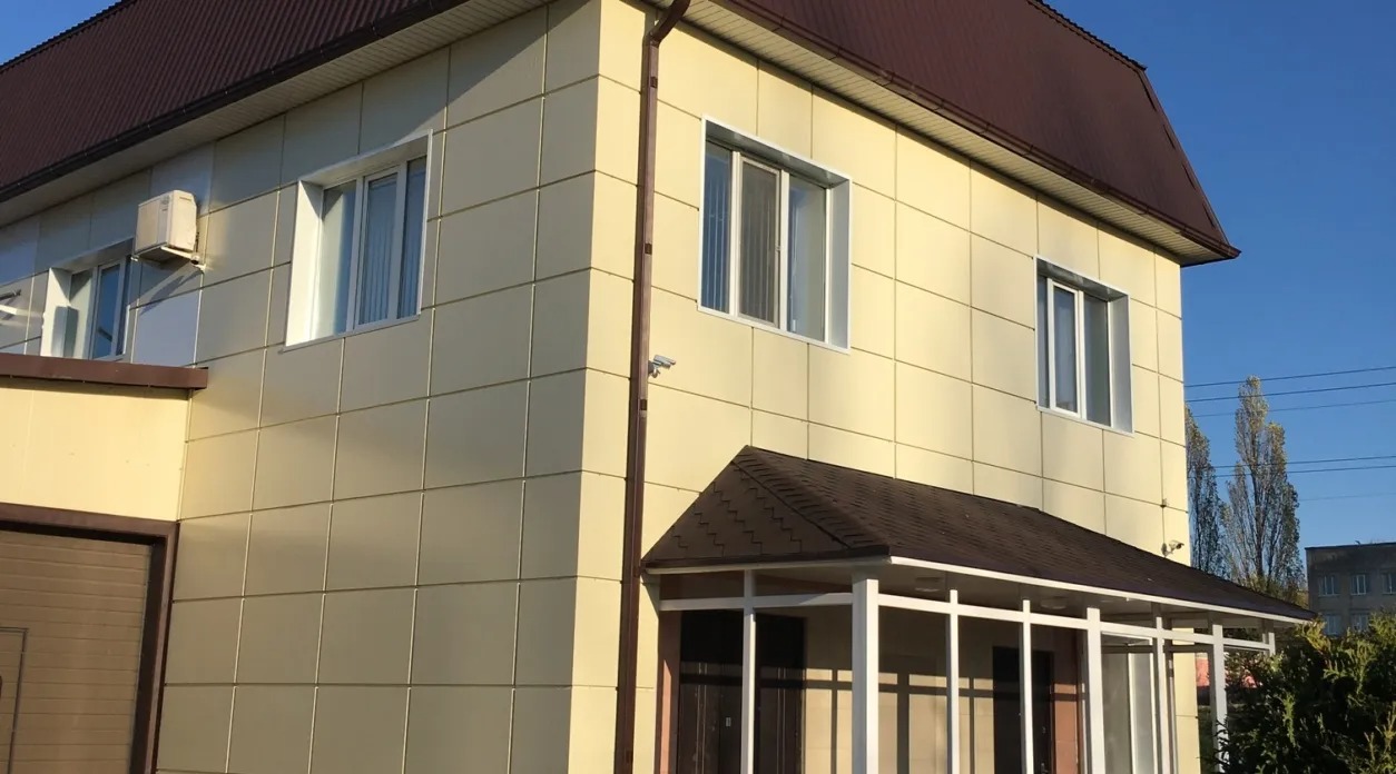 Żółta elewacja budynku z brązowym dachem i białymi oknami, widoczny klimatyzator i kamera monitoringu.