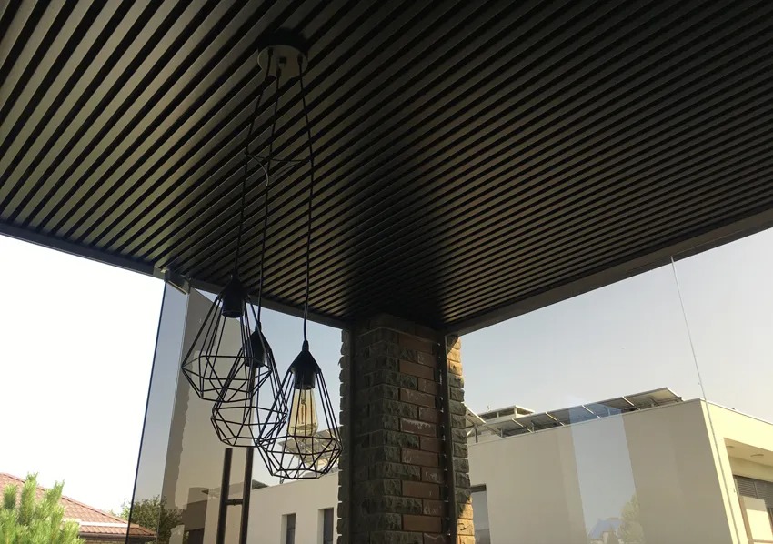 Zadaszenie tarasu z lamelami w kolorze ciemnego brązu, oświetlone trzema lampami wiszącymi w stylu industrialnym z żarówkami Edisona. Widoczny ceglany filar i szklana ściana tarasu.
