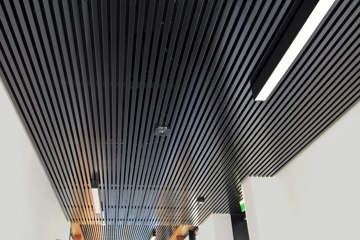 Nowoczesny sufit rastrowy z czarnych, pionowych lameli, oświetlenie liniowe LED, widoczne elementy instalacji.