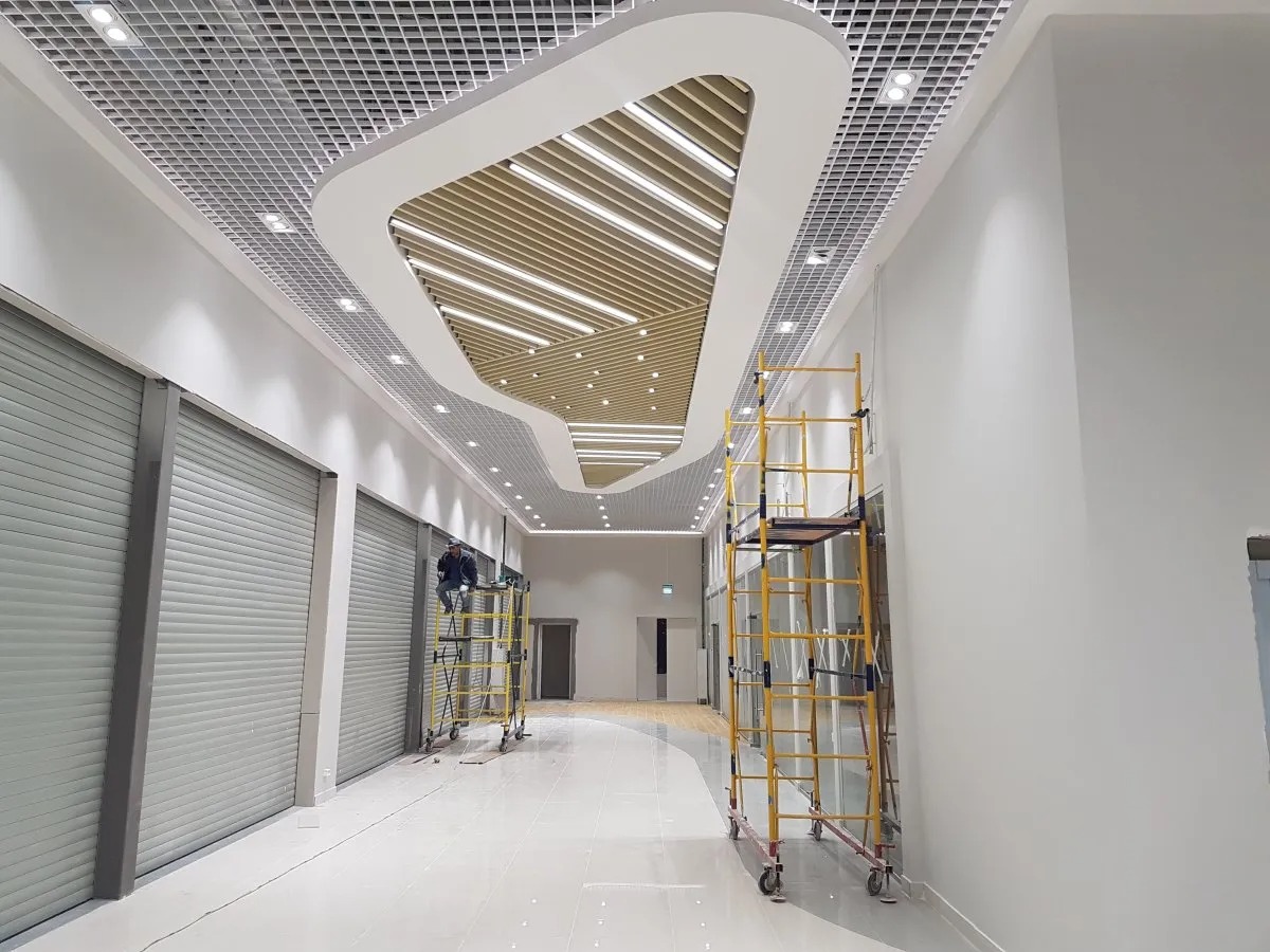 Wnętrze komercyjne w trakcie wykańczania: widok na sufit z dekoracyjnymi elementami i siatką, pracownik na rusztowaniu przy roletach oraz żółte rusztowanie na błyszczącej podłodze.