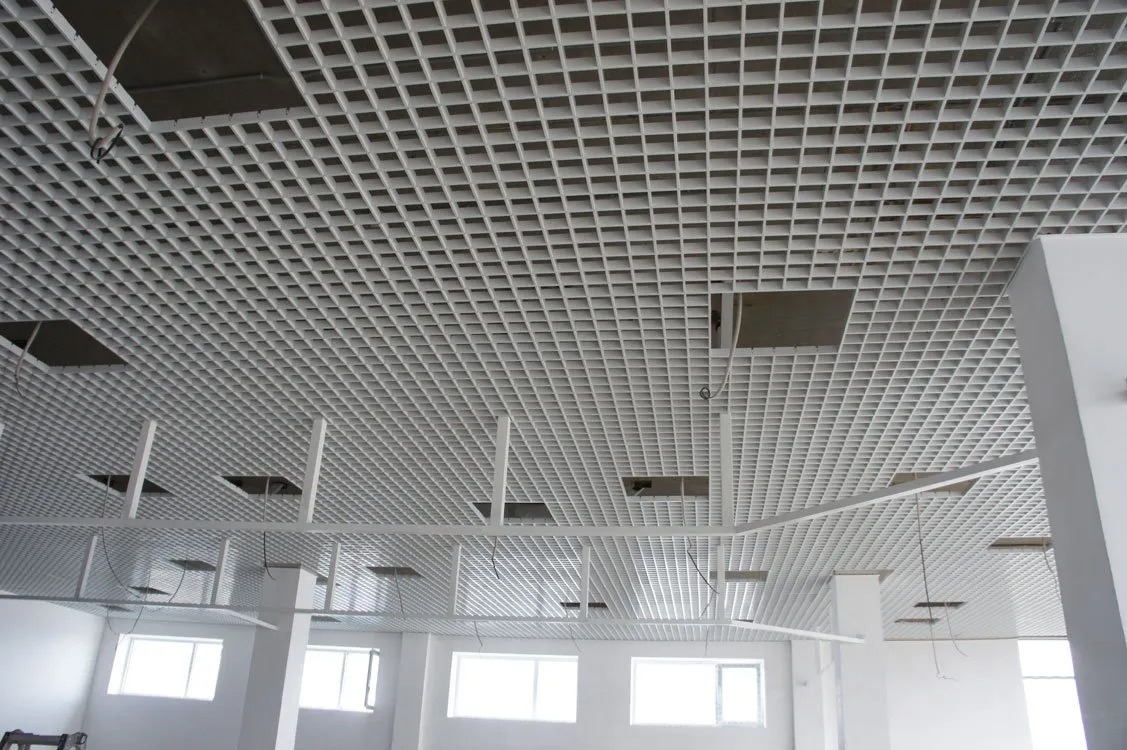 Biały sufit rastrowy z kwadratowymi oczkami w przestrzeni biurowej, widoczne elementy instalacji i niedokończone fragmenty.