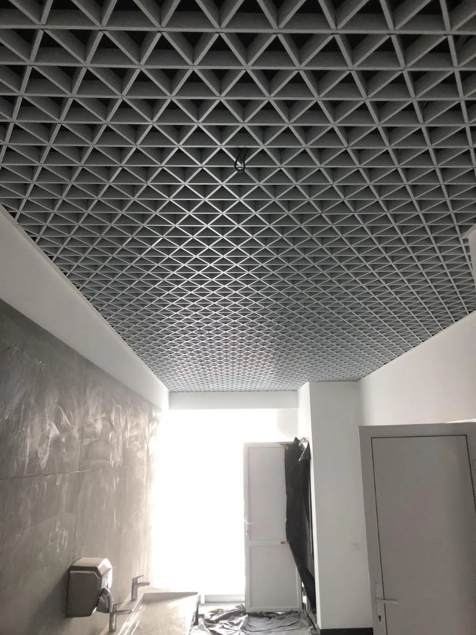 Sufit rastrowy o trójkątnej strukturze w pomieszczeniu z surową ścianą i otwartymi drzwiami na końcu korytarza.