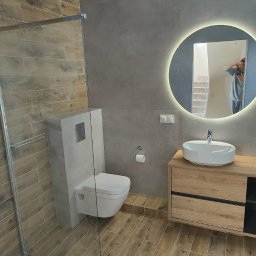 BUDCLEAN SPÓŁKA Z OGRANICZONĄ ODPOWIEDZIALNOŚCIĄ - Nowoczesna łazienka z kabiną prysznicową bez brodzika, podwieszaną toaletą w szarej obudowie i okrągłym lustrem z podświetleniem LED, odbijającym schody w tle.