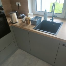 BUDCLEAN SPÓŁKA Z OGRANICZONĄ ODPOWIEDZIALNOŚCIĄ - Szary zlew wkomponowany w drewniany blat w nowej kuchni, obok czajnik elektryczny i drobne akcesoria kuchenne, widok na okno z zielenią na zewnątrz.