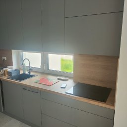 BUDCLEAN SPÓŁKA Z OGRANICZONĄ ODPOWIEDZIALNOŚCIĄ - Szare szafki kuchenne z blatem w kolorze drewna, czarnym zlewem i płytą indukcyjną, widok na okno z widokiem na zieleń na zewnątrz.