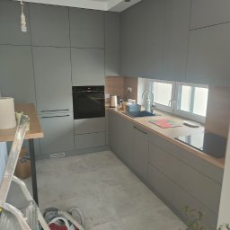 BUDCLEAN SPÓŁKA Z OGRANICZONĄ ODPOWIEDZIALNOŚCIĄ - Szara kuchnia z zabudową meblową pod sufit, widoczny piekarnik, blat z jasnego drewna, okno oraz drabina i odkurzacz w trakcie prac wykończeniowych.