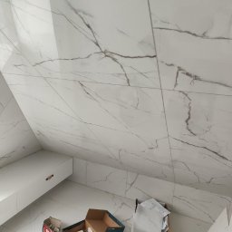 BUDCLEAN SPÓŁKA Z OGRANICZONĄ ODPOWIEDZIALNOŚCIĄ - Ukośna ściana i podłoga wyłożone białymi, marmurkowymi płytkami, częściowo widoczna biała szafka wisząca i kartony z akcesoriami glazurniczymi na podłodze.