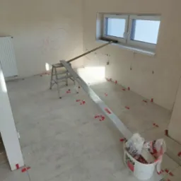 Układanie szarych płytek podłogowych w jasnym pomieszczeniu, widoczne poziomice, drabina, wiadro z akcesoriami i czerwone kliny dystansowe, na ścianie rysunek kredą.