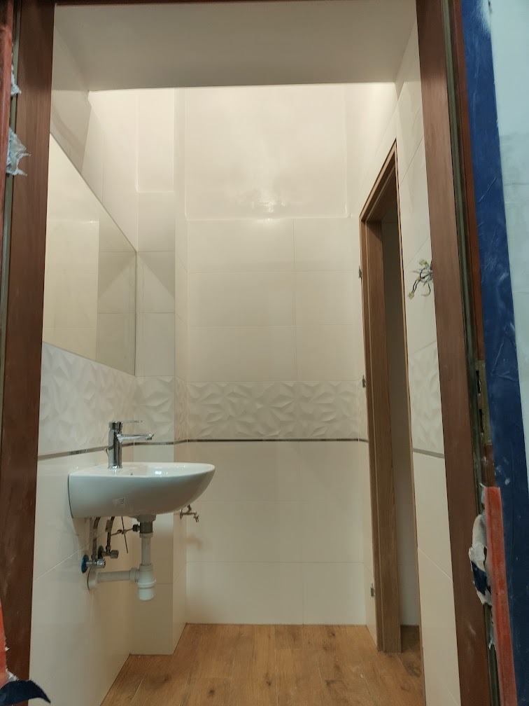 Mała łazienka w trakcie wykańczania: białe płytki z dekoracyjnym pasem, umywalka zamontowana na ścianie, drewnopodobna podłoga, widoczne niedokończone obramowanie drzwi.