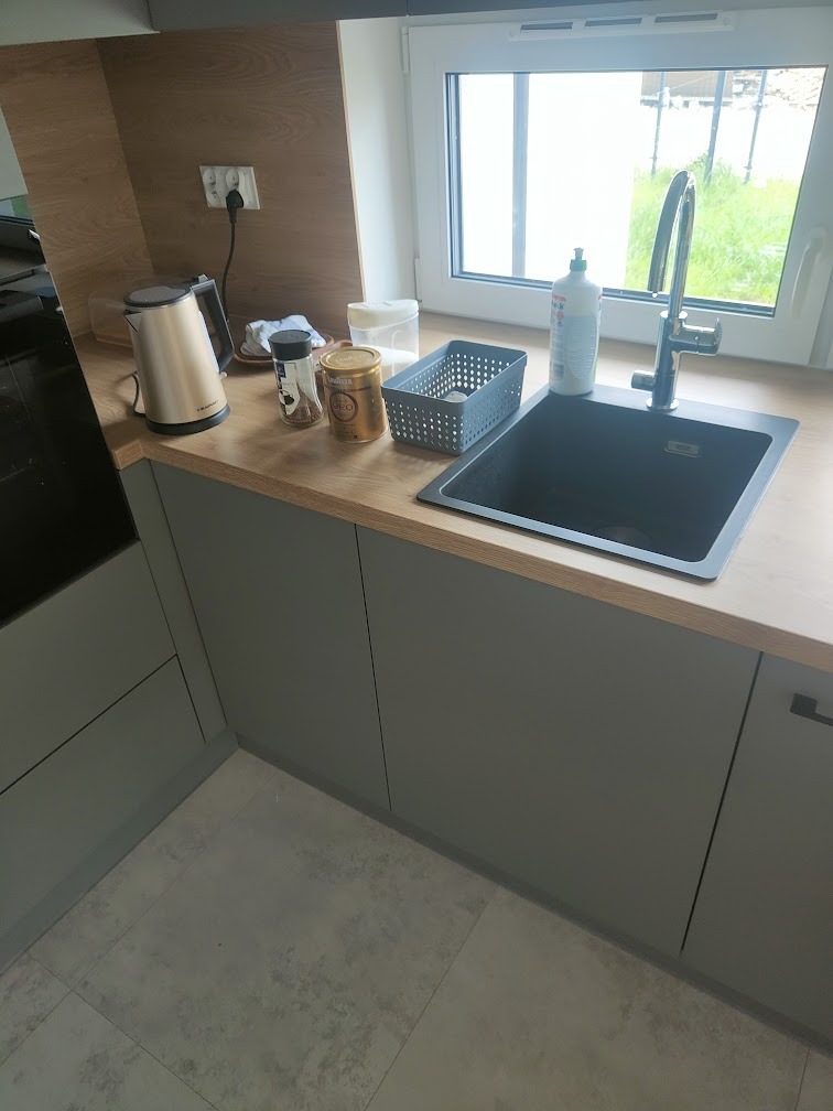 Szary zlew wkomponowany w drewniany blat w nowej kuchni, obok czajnik elektryczny i drobne akcesoria kuchenne, widok na okno z zielenią na zewnątrz.