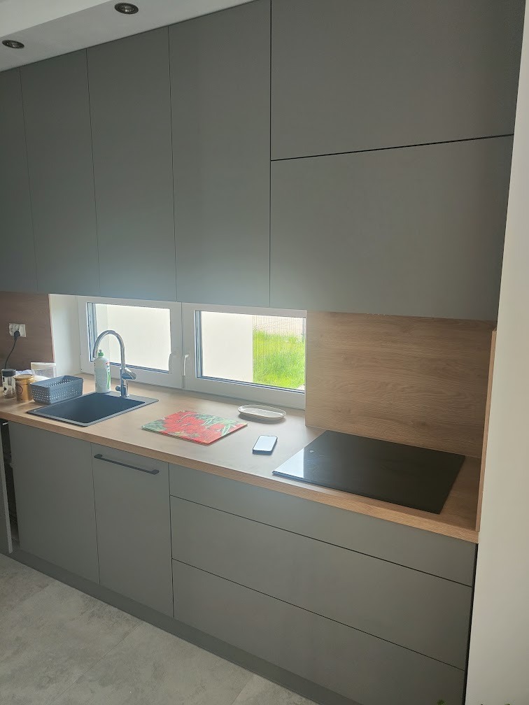 Szare szafki kuchenne z blatem w kolorze drewna, czarnym zlewem i płytą indukcyjną, widok na okno z widokiem na zieleń na zewnątrz.