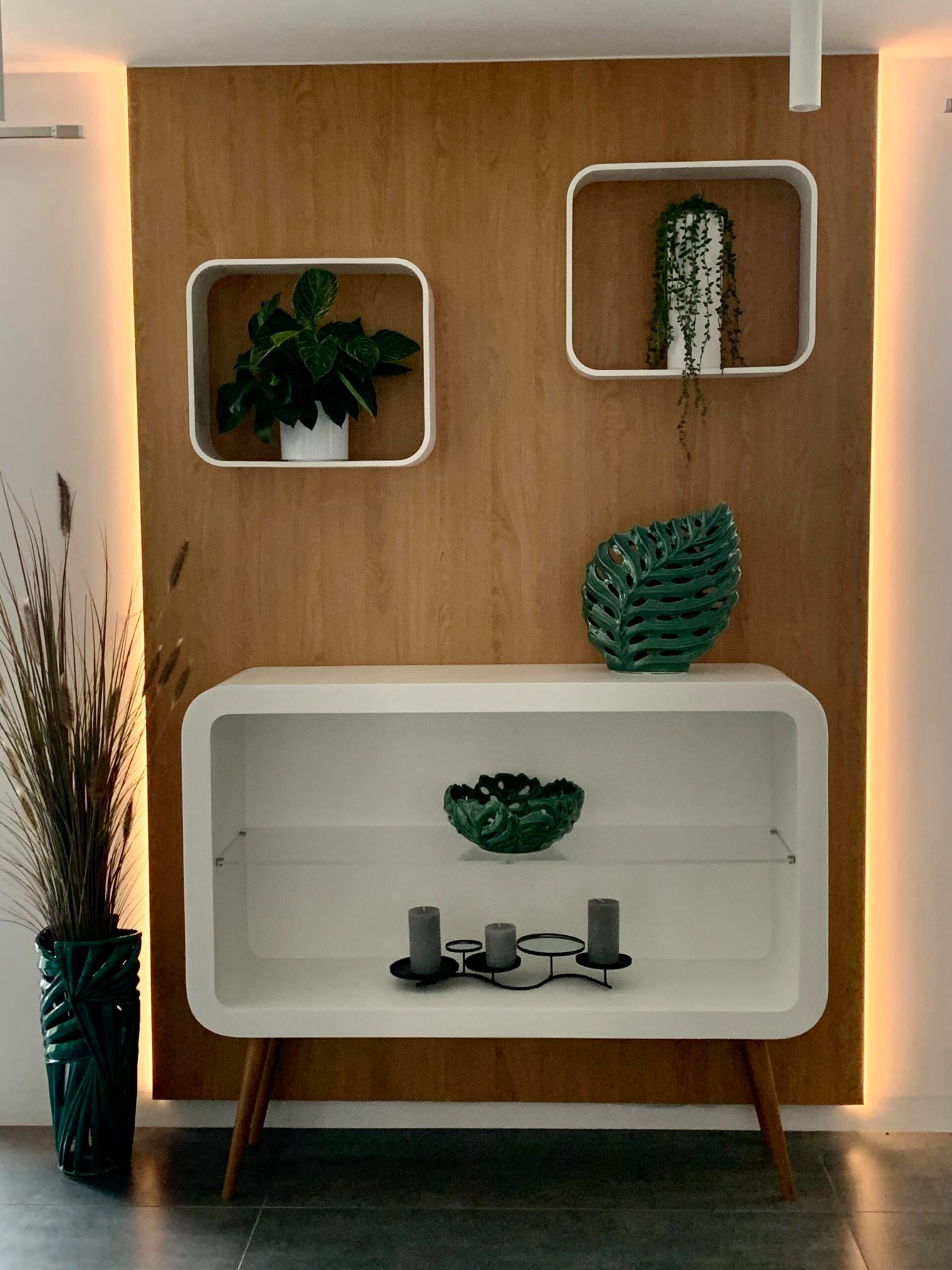 Nowoczesny biały kredens na drewnianych nóżkach, z dekoracjami: rośliny w kwadratowych ramkach, ceramiczna misa i liść monstery, oraz świece w minimalistycznym świeczniku.