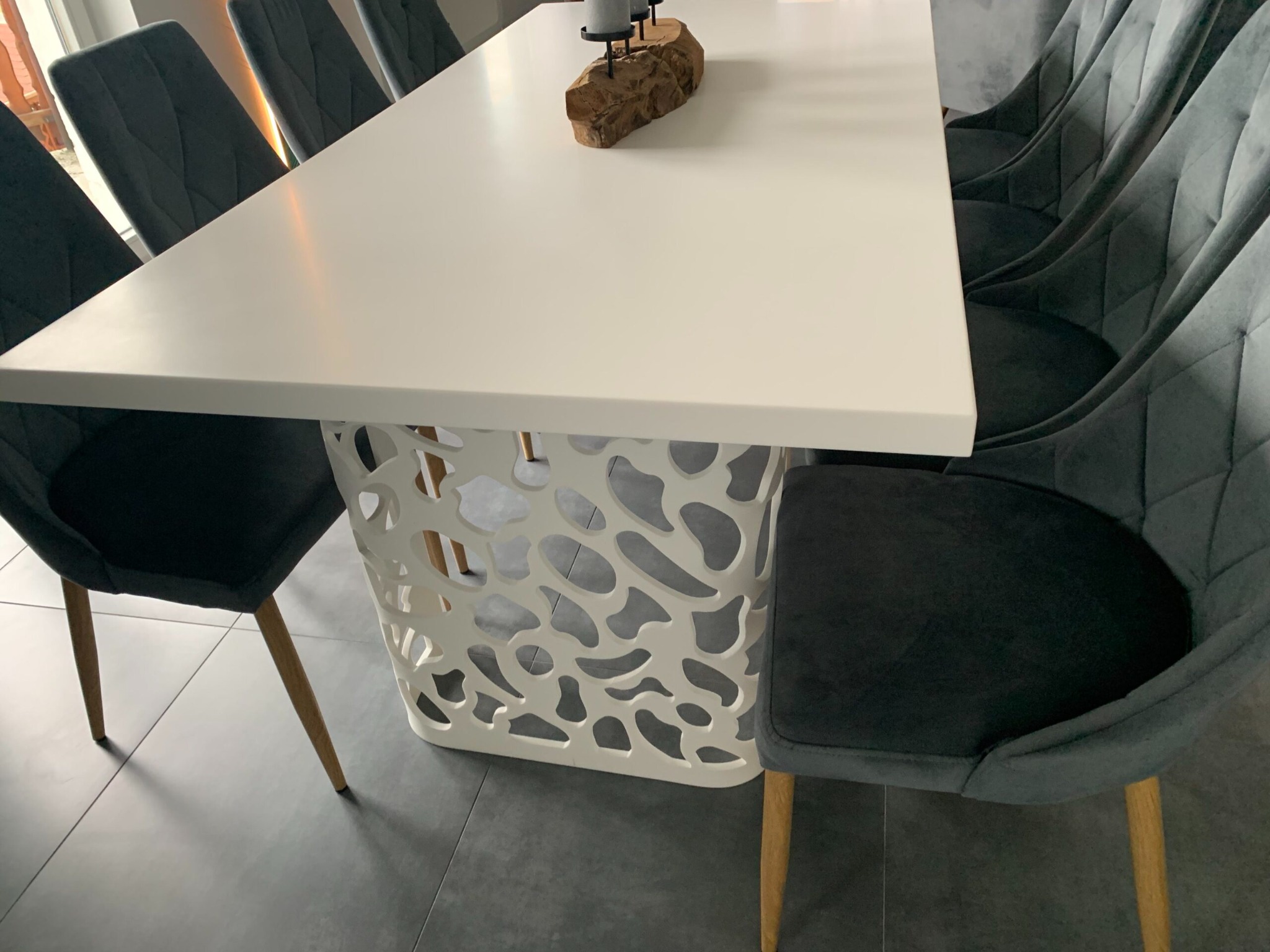 Nowoczesny stół z białym blatem i ażurową podstawą, otoczony szarymi krzesłami z pikowanymi oparciami, na szarej podłodze.