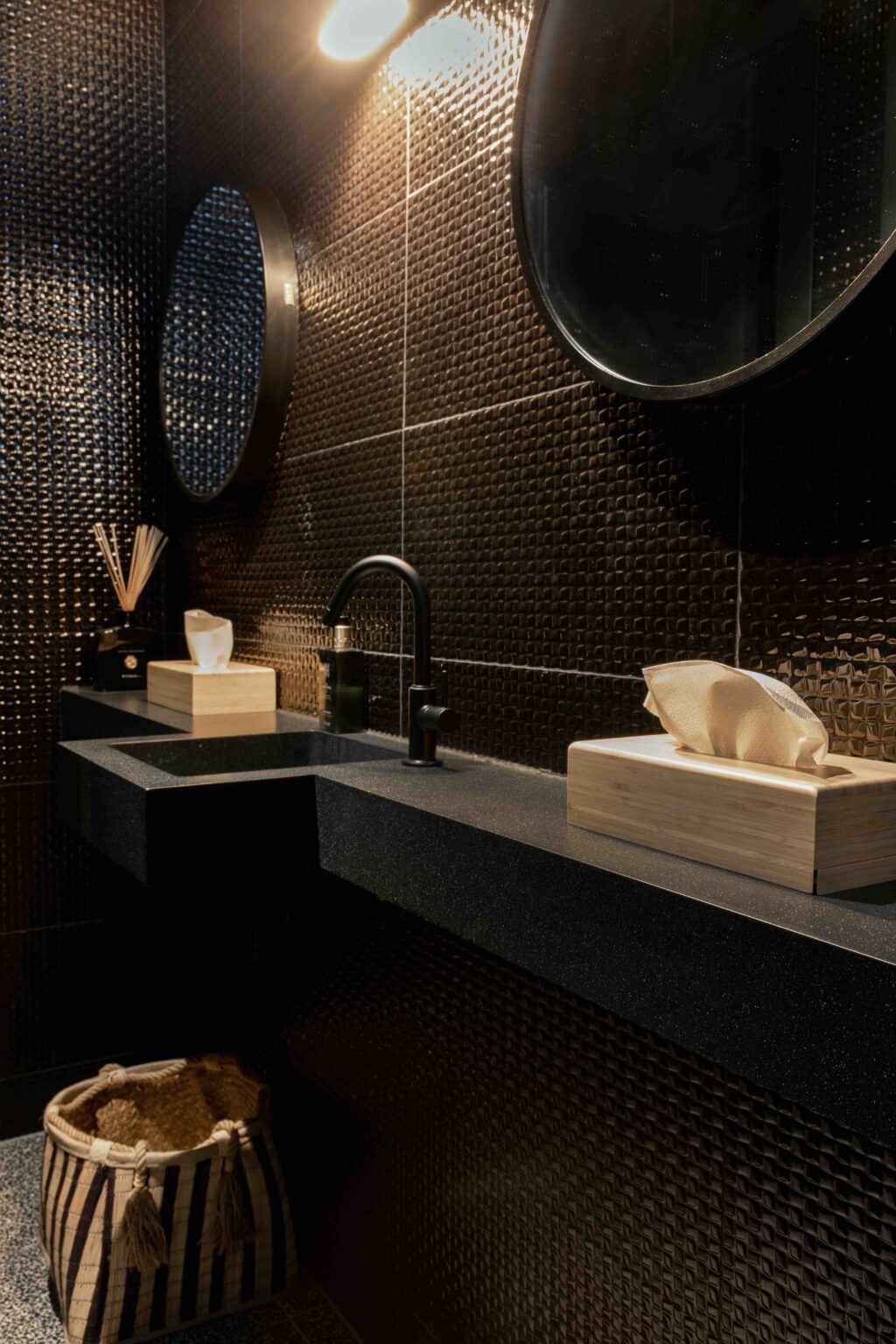 Czarna, minimalistyczna łazienka z umywalką nablatową, czarną baterią i teksturowanymi, ciemnymi płytkami na ścianie. Na blacie z widoczną fakturą stoi drewniane pudełko na chusteczki i pojemnik...