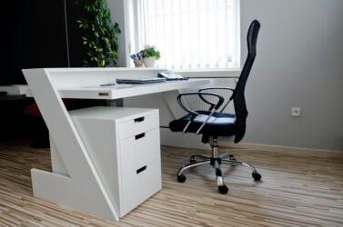 Nowoczesne białe biurko z szufladami i charakterystyczną ukośną nogą, z czarnym krzesłem biurowym na kółkach, na tle szarej ściany i okna z rośliną.