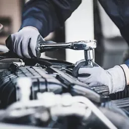 Otwarta maska samochodu z widocznym silnikiem, nad którym pracują ręce w szarych rękawicach, używając klucza nasadowego.