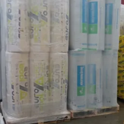 Pakiety wełny mineralnej Eurowool i Knauf Insulation, zabezpieczone folią i umieszczone na paletach, oczekują na transport w Ostrowi Mazowieckiej. Wilgotna posadzka sugeruje niedawne opady.