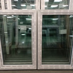 e-oknadrzwi.pl Materiały Budowlane Online - Białe ramy okienne z PCV, ułożone w stos, widoczne szyby z odblaskami magazynowego oświetlenia.