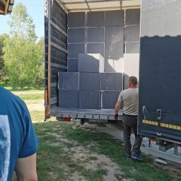 e-oknadrzwi.pl Materiały Budowlane Online - Rozładunek szarych bloków ociepleniowych z naczepy ciężarówki przez pracownika w rękawicach roboczych, widoczne tablice rejestracyjne pojazdu.