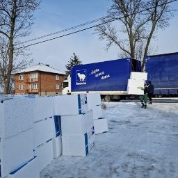 e-oknadrzwi.pl Materiały Budowlane Online - Białe bloki materiału izolacyjnego ułożone na śniegu przed ciężarówką z napisem 'ciepła strona domu' i logiem niedźwiedzia polarnego, w tle budynek mieszkalny i drzewa.