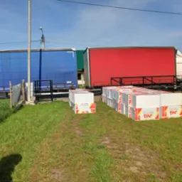 Dwa samochody ciężarowe z naczepami w kolorze niebieskim i czerwonym zaparkowane obok siebie. Na trawie ułożone palety z białymi paczkami materiału budowlanego z czerwono-żółtymi oznaczeniami.