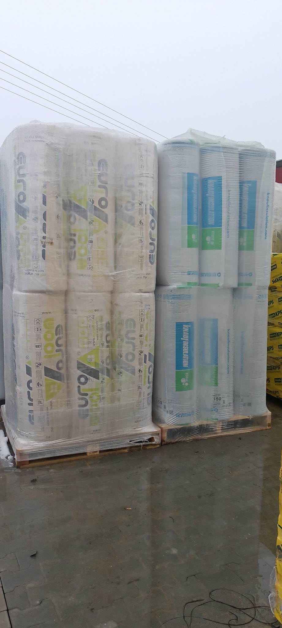 Pakiety wełny mineralnej Eurowool i Knauf Insulation, zabezpieczone folią i umieszczone na paletach, oczekują na transport w Ostrowi Mazowieckiej. Wilgotna posadzka sugeruje niedawne opady.