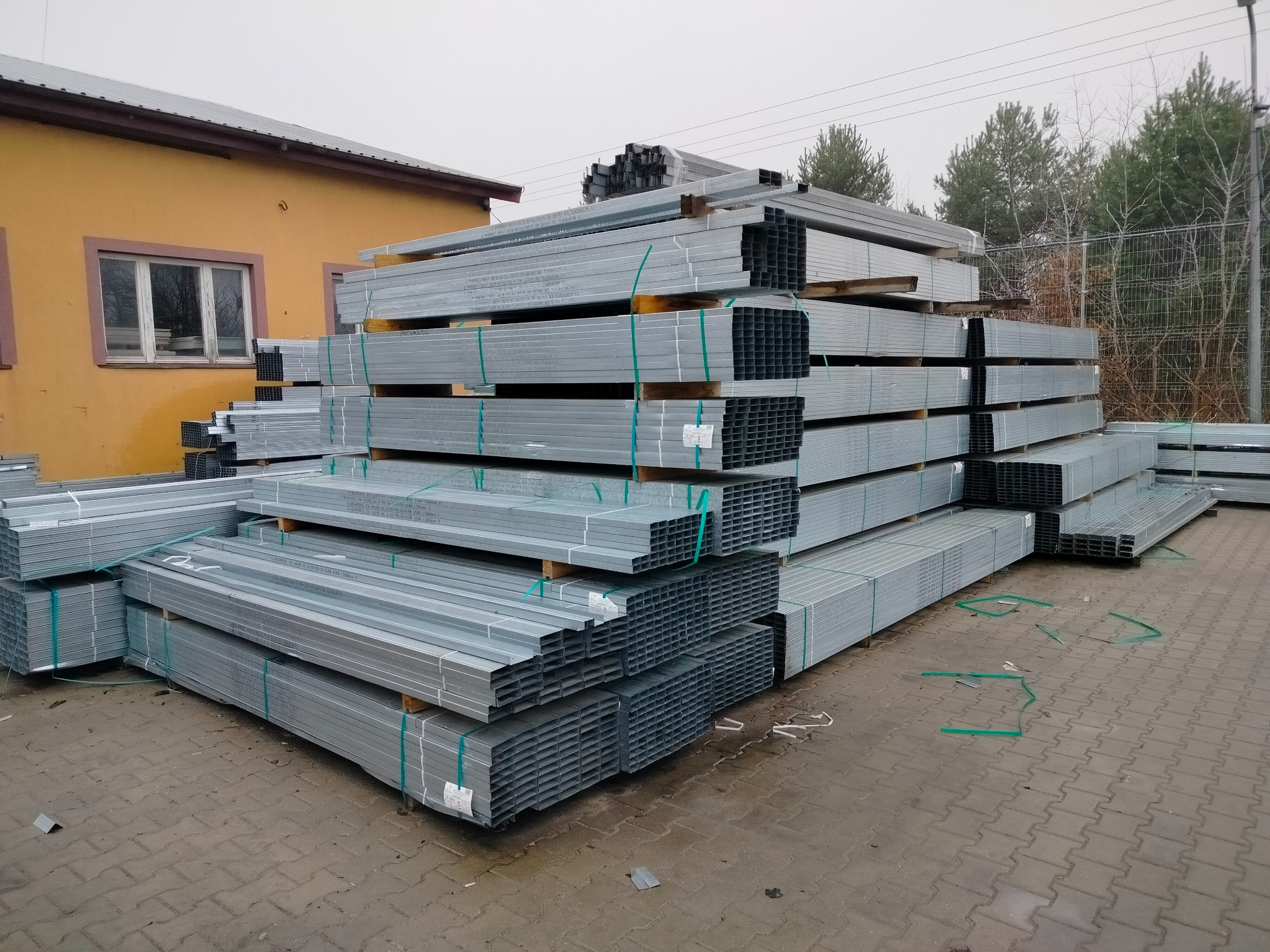 Stosy ocynkowanych profili stalowych o przekroju kwadratowym, ułożone na paletach, zabezpieczone zielonymi taśmami, na placu budowlanym w pochmurny dzień.